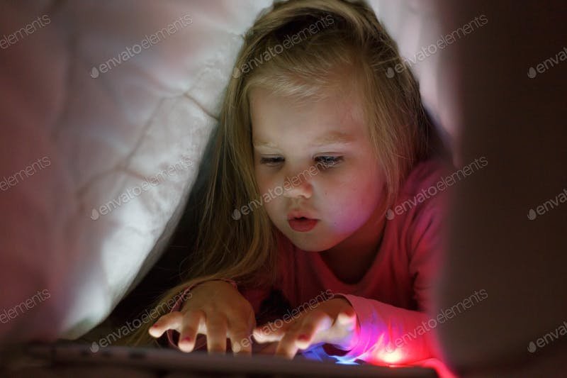 A Little Girl Playing On Her Tablet Under The Blanket PSD Template.