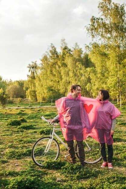 Vertical shot of a couple sharing a pink plastic raincoat at a date with a bike Free Photo